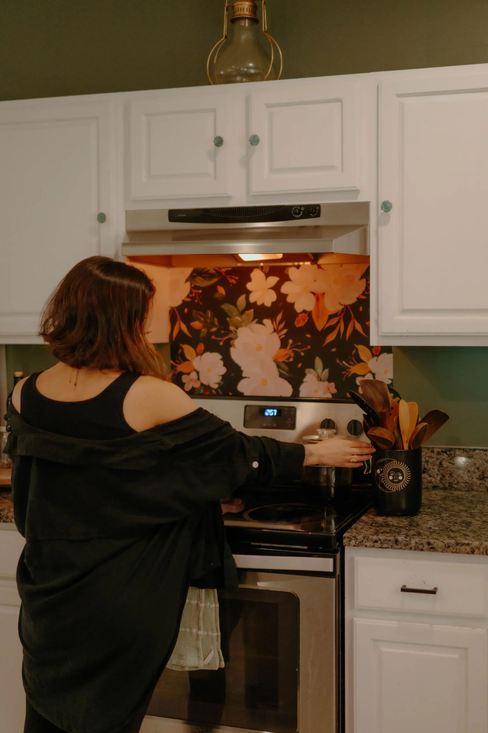 woman wearing black in front of stove with floral wallpaper backsplash in green and white kitchen